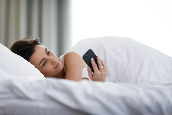 美しいです若いです女性睡眠使用彼女のスマートフォンでベッド最初事で朝 — ストック写真