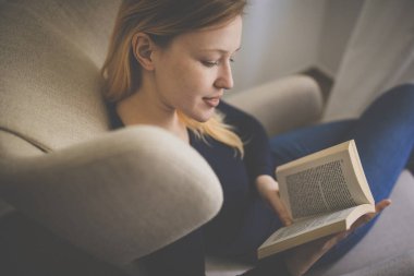 Tasarımcı koltuğunda kitap okuyan sevimli genç bir kadın. Renkli resim, sığ DOF.