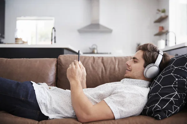 携帯電話から自宅のソファーに横になっている男を着て無線ヘッドフォンのストリーミング — ストック写真