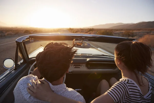 Rear View Of Couple On Road Trip Driving Classic Convertible Car Towards Sunset