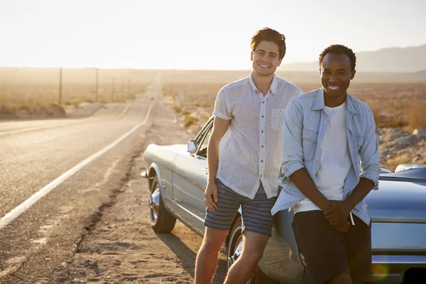 Two Male Friends Enjoying Road Trip Standing Next To Classic Car On Desert Highway