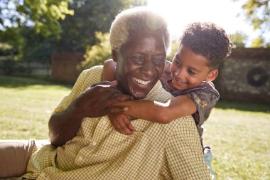 Senior black man sitting on grass, embraced by his grandson clipart