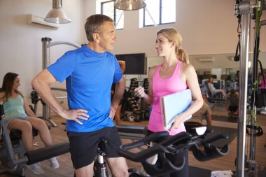 Senior Man Exercising On Cycling Machine Being Encouraged By Female Personal Trainer In Gym clipart