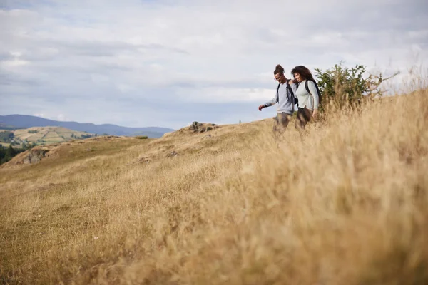 混合种族年轻的成年夫妇微笑着 而在山间徒步时穿过下坡的田野 — 图库照片