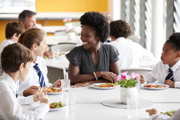 女教師制服テーブルの周りに座って カフェテリアでランチを食べることを身に着けている高校生のグループに — ストック写真