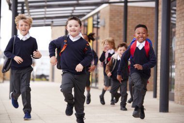 Excited primary school kids, wearing school uniforms and backpacks, running on a walkway outside their school building, front view, close up clipart