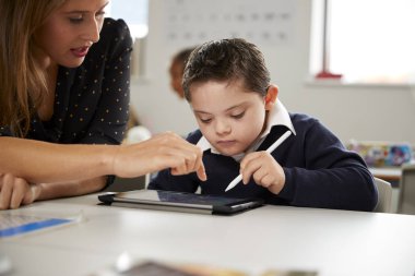 Young female teacher working with a Down syndrome schoolboy sitting at desk using a tablet computer in a primary school classroom, front view, close up clipart