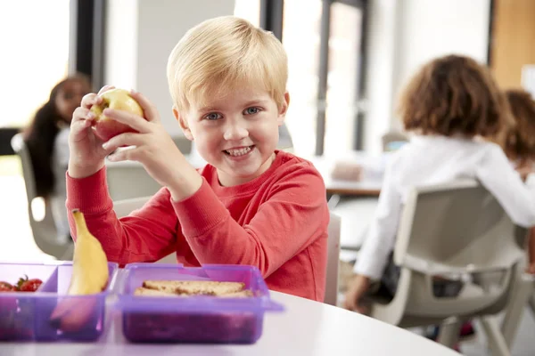 昼食休憩中に幼稚園の教室でリンゴを笑顔で抱きしめてテーブルに座る若い白人男子学生 — ストック写真