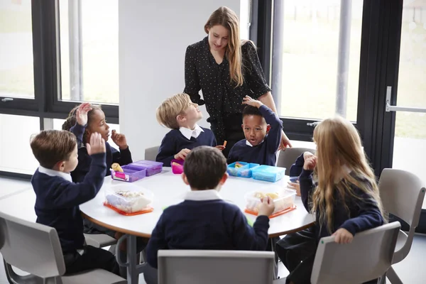 女性教師は お弁当を食べながら丸いテーブルで一緒に座っている小学生のグループに話しかける — ストック写真