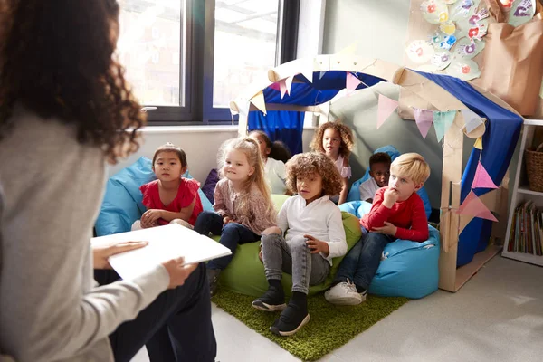 教室の快適なコーナーで豆袋の上に座っている子供たちのグループに本を読んで椅子に座っている女性の幼児学校の教師は 高い眺め クローズアップ — ストック写真
