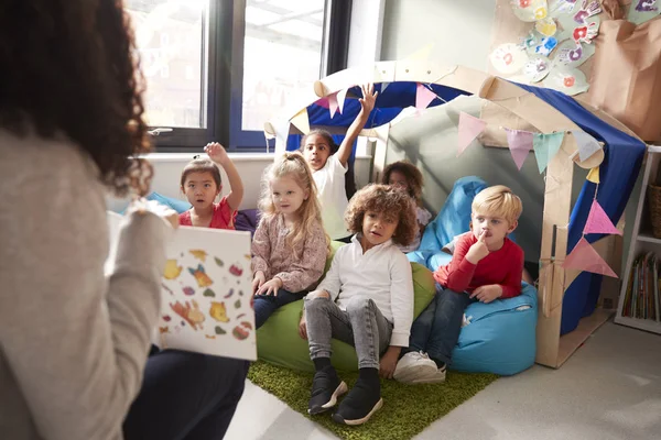 女幼儿学校老师坐在椅子上向坐在豆袋上的一群孩子展示一本书 — 图库照片