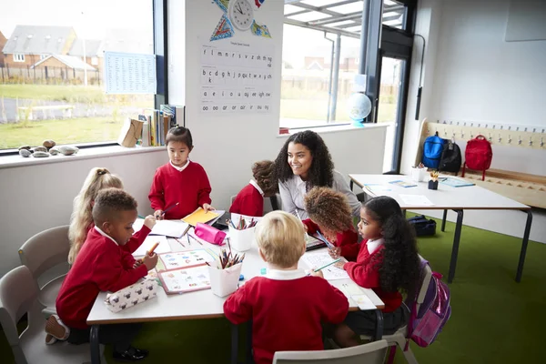 A group of infant school kids sitting at a table in a classroom with their female teacher Stock Picture