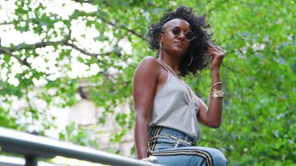 トレンディな若い黒人女性着用キャミソール サイド ストライプ ジーンズ サングラス 髪の毛で遊んで手すりの上に座って低い角度 — ストック動画