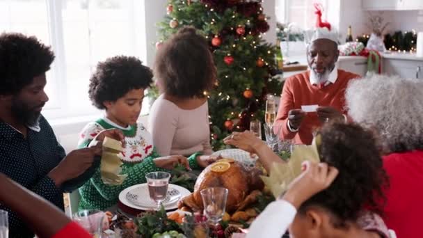 多世代家族はジョークを読んで クリスマス クラッカーから紙のパーティの帽子に置くことのクリスマスの夕食のテーブルに座って — ストック動画