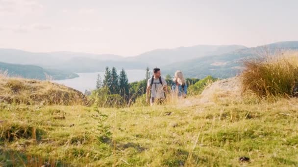 微笑的年轻夫妇穿着背包爬山徒步穿越屋湖区与美丽的风景背景 拍摄慢镜头 — 图库视频影像