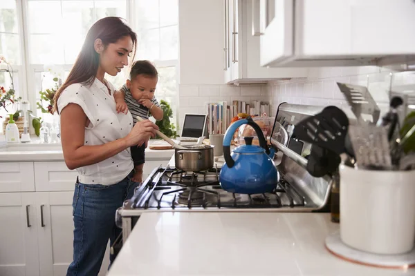 多任务妈妈抱着她的宝宝 同时在厨房的炉灶 侧视图做食物 — 图库照片