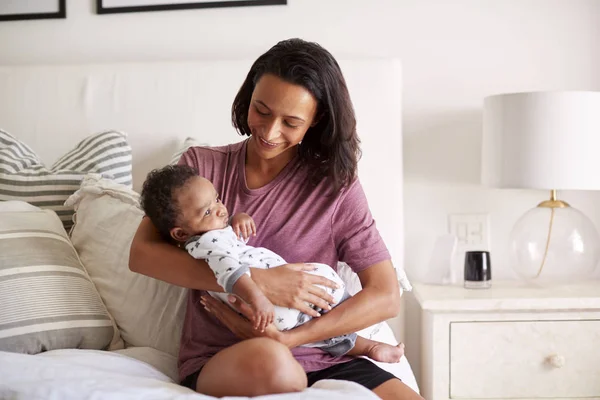 ミレニアル世代の混合レースの母親は彼女の腕の中で彼女の3ヶ月の赤ちゃんの息子を保持して彼女のベッドに座って — ストック写真