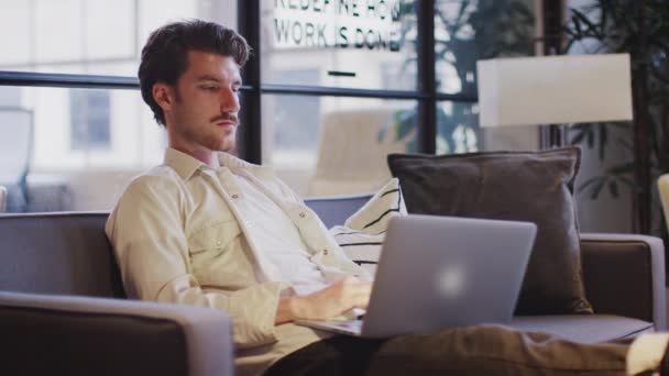 オフィスラウンジエリアで足を上げて座っているラップトップで働く若い男 側面の景色 — ストック動画