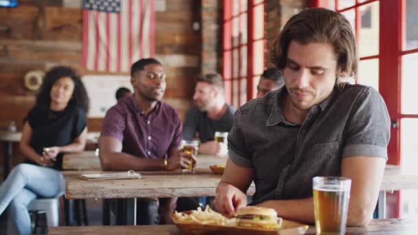 ハンバーガーやフライドポテトを食べながらゲームを見てスポーツバーに座っている男 スローモーションで撮影 — ストック動画