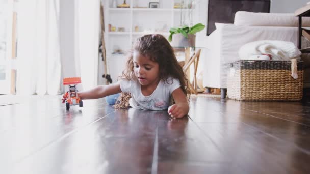おもちゃの掘削トラックで遊んで居間の床に横たわっている若いヒスパニックの女の子 正面図 — ストック動画