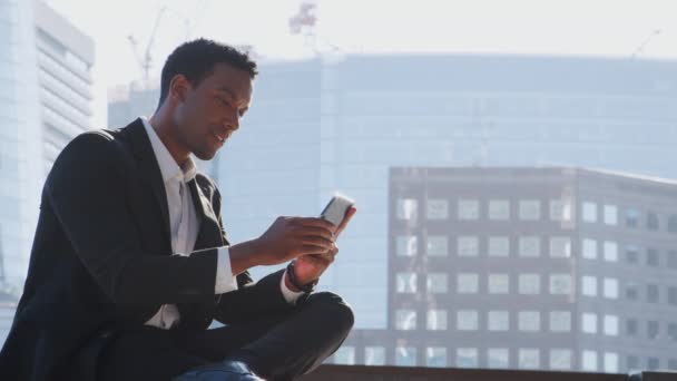 千禧年黑人商人穿着西装和白色衬衫坐在泰晤士河堤上用他的智能手机 — 图库视频影像