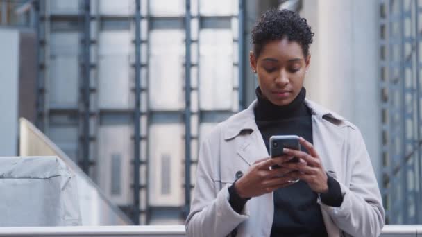 彼女のスマートフォン フロントビュー ウエストアップを使用して 近代的な建物の前に街に立っているファッショナブルな若い黒人女性のクローズアップ — ストック動画