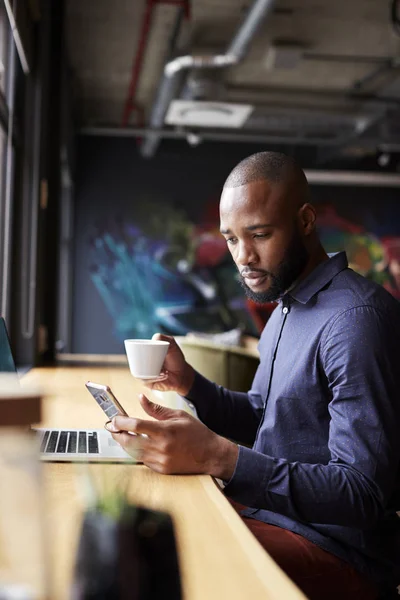 ミッドアダルト黒人男性クリエイティブは スマートフォン サイドビュー 垂直を使用してコーヒーを持つ窓のそばに座って — ストック写真