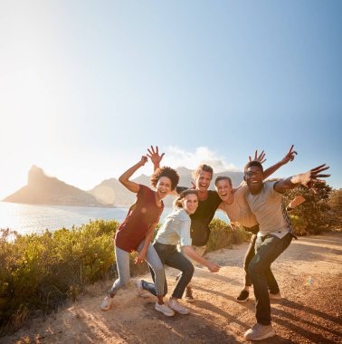 Five millennial friends on a road trip have fun posing for photos on a coastal path, full length clipart