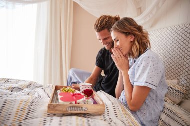 Millennial white man surprising his female partner with breakfast and gifts in bed, close up clipart