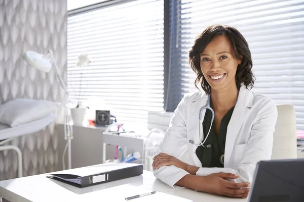 微笑的女医生的肖像穿着白色外套与听诊器坐在办公桌后面在办公室 — 图库照片