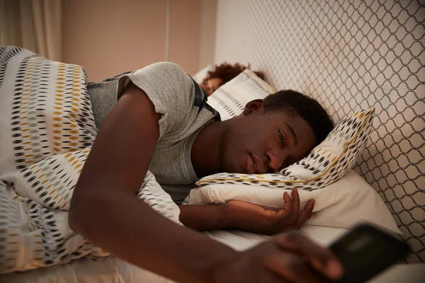 ミレニアルアフリカ系アメリカ人男性は スマートフォンを持ってベッドで半分眠り バックグラウンドで彼のパートナー クローズアップ — ストック写真