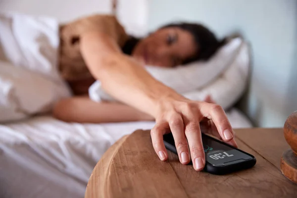 ベッドに横たわる中年の大人の女性は 前景のベッドサイドテーブルの上でスマートフォンに手を差し伸べる — ストック写真