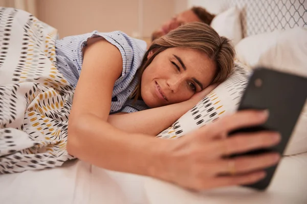 Mulher Branca Meio Dormindo Cama Olhando Para Smartphone Parceiro Dormindo — Fotografia de Stock