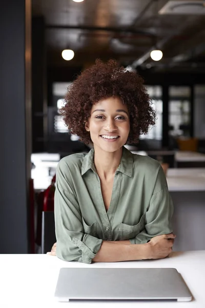 千禧年黑人女性创意坐在办公桌在一个开放的计划办公室微笑的相机 — 图库照片