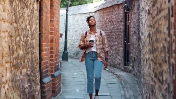 年轻女子穿着格子衬衫和蓝色牛仔裤走向相机在老石墙之间的小巷拿着外卖咖啡 选择性的焦点 — 图库视频影像