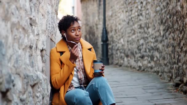 黄色いエンドウ豆のコートと青いジーンズを着た若い女性は イヤホンを使ってスマートフォンで話している歴史的な都市の路地に座って クローズアップ — ストック動画