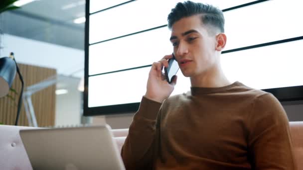 カジュアルなオフィスのソファに座ってラップトップコンピュータで作業する携帯電話を使用して若いビジネスマンは 閉じる 低い角度 — ストック動画