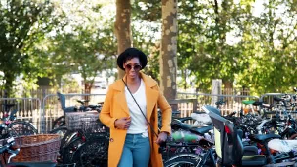サングラス 青いジーンズ ボタンのない黄色いエンドウ豆のコートと駐車中の自転車の中を歩くクロスボディのハンドバッグを着て カメラに微笑むファッショナブルな若い黒人女性 — ストック動画
