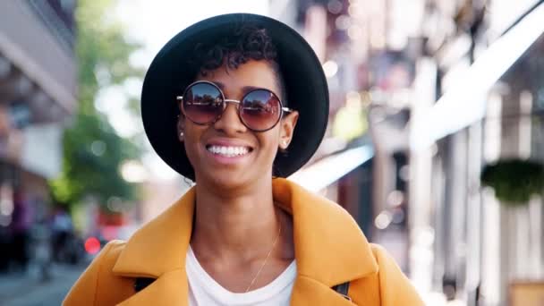 黄色いエンドウ豆のコート サングラス ホンブルクの帽子をかぶったミレニアル世代の黒人女性が カメラに微笑みかかって街の通りに立ち クローズアップ — ストック動画