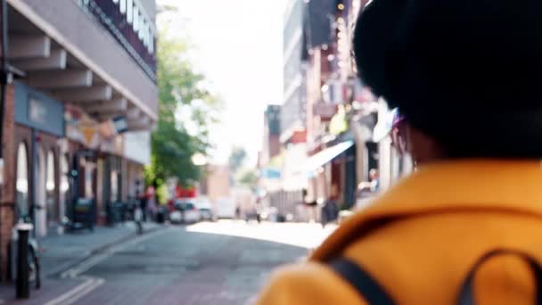 帽子と黄色いエンドウ豆のコートを着て通りを歩き カメラに振り返るファッショナブルな若い黒人女性のバックビュー — ストック動画