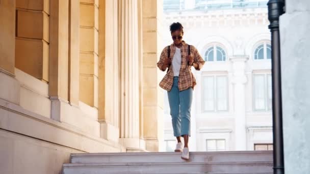 年轻的黑人妇女穿着格子衬衫和蓝色牛仔裤走下街道楼梯在历史建筑的入口 — 图库视频影像