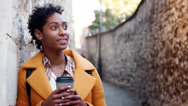 テイクアウトコーヒーを飲む路地で石垣に寄りかかって黄色いコートを着たミレニアルの黒人女性 前景に焦点を当てる — ストック動画