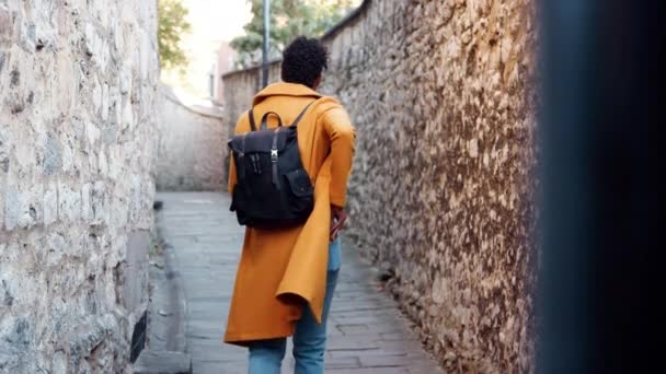 黄色いエンドウ豆のコートを着た若い黒人女性がジーンズの後ろポケットにスマートフォンを入れ 石垣の間の狭い路地をカメラから歩き去る 選択的な焦点 — ストック動画