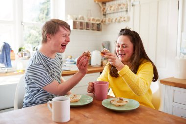Young Downs Syndrome Couple Enjoying Tea And Cake In Kitchen At Home clipart
