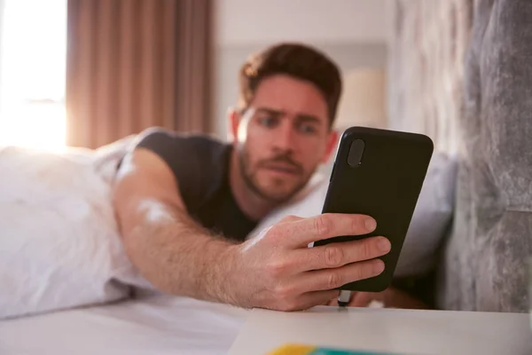 ベッドで目を覚ます男はすぐに携帯電話を見るために手を伸ばす — ストック写真