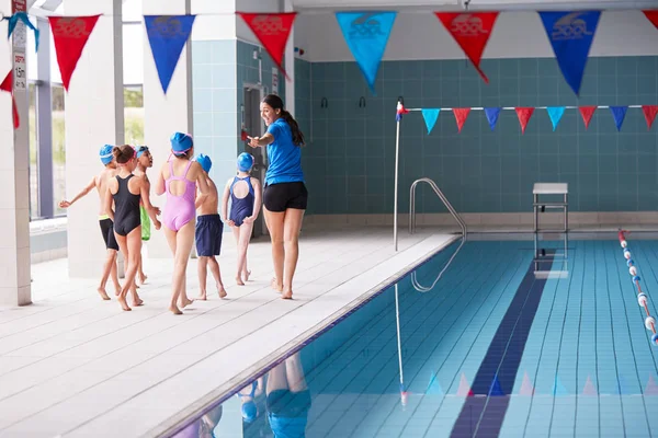 Female Coach Walks Children In Swimming Class Along Edge Of Indoor Pool