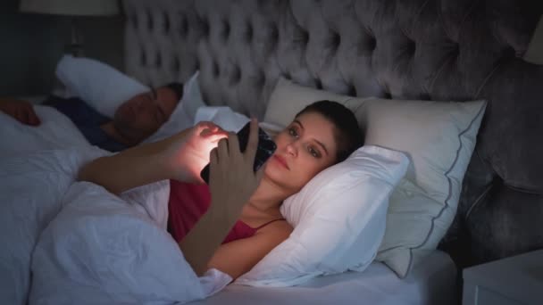 Woman Lying Bed Checking Mobile Phone Whilst Male Partner Sleeps — Stock Video