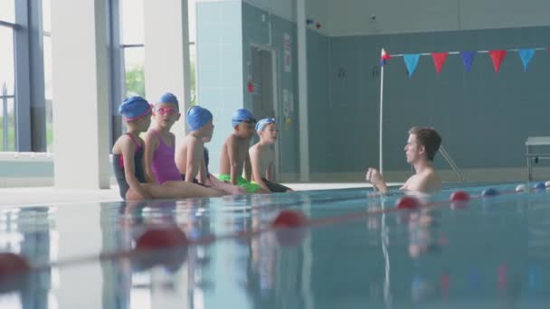 水中の男性コーチは 水泳クラスの屋内プールの指示の端に座っている子供たちを与えます スローモーションで撮影 — ストック動画