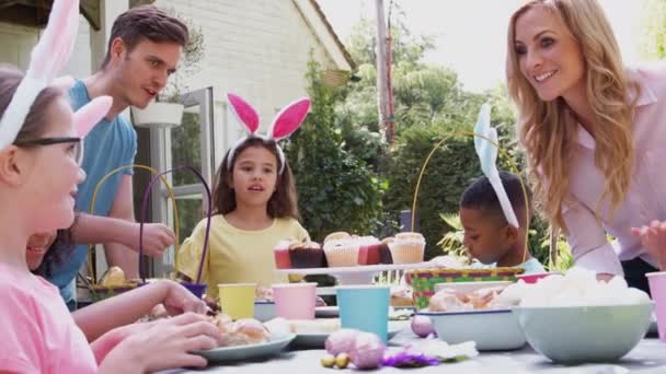 親と一緒にイースターパーティーを楽しむ屋外のテーブルに座っているウサギの耳を身に着けている子供たちのグループ スローモーションで撮影 — ストック動画