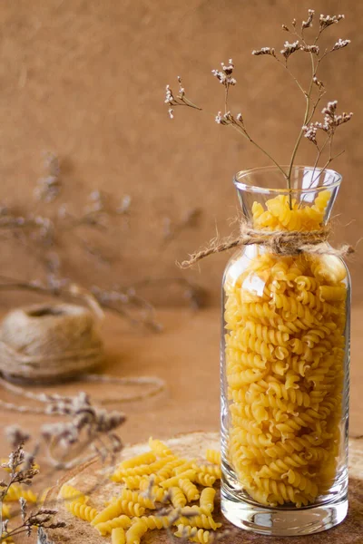 Spiral pasta in a glass jar on a wooden stand, next to it are dry tree branches, on a beige background